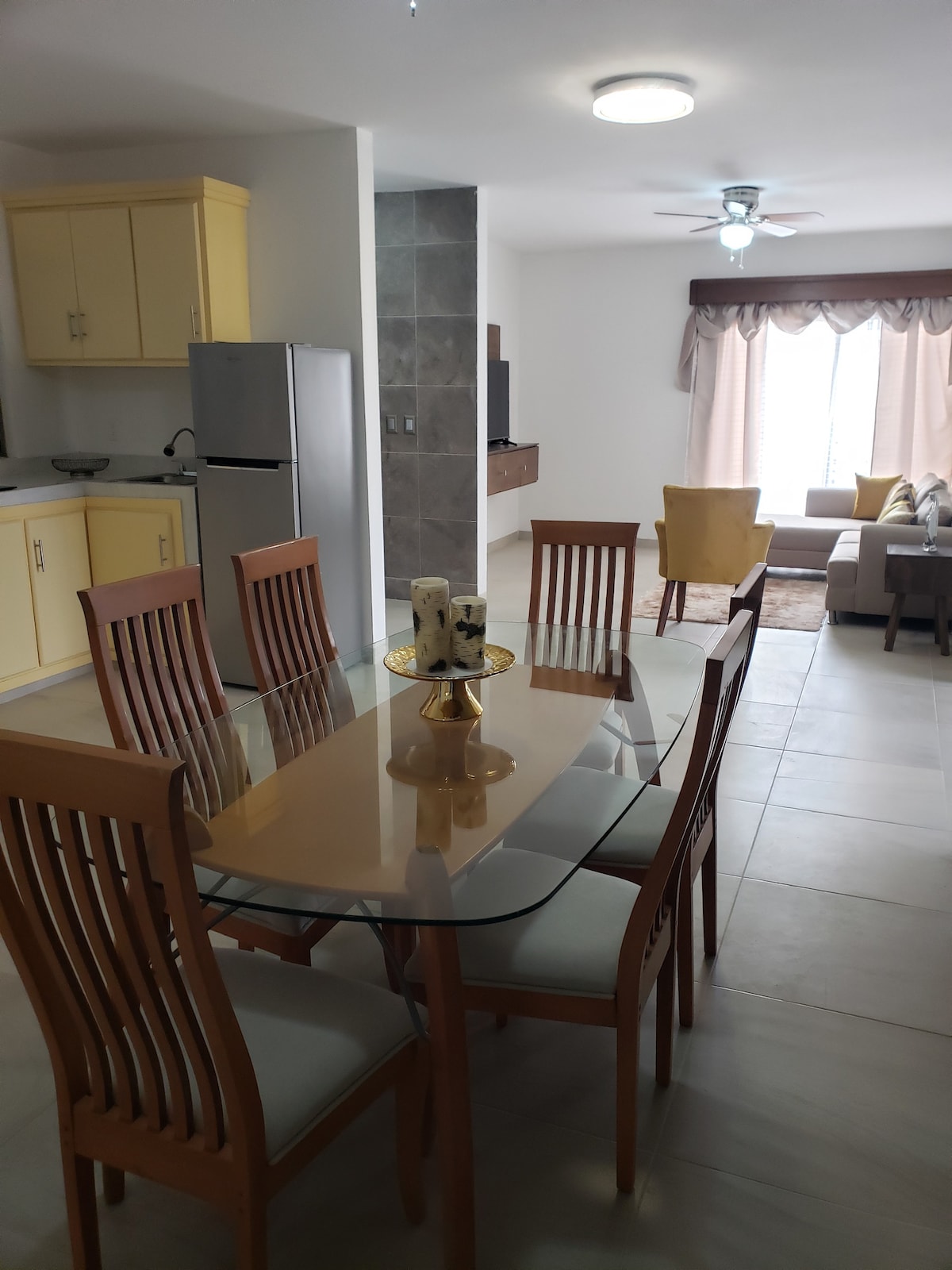 A dining area is placed adjacent to a modern kitchen, featuring a rectangular glass table surrounded by six wooden chairs. A cozy living area is visible, showcasing a light-colored sofa and window drapes. Natural light fills the space through the large window.