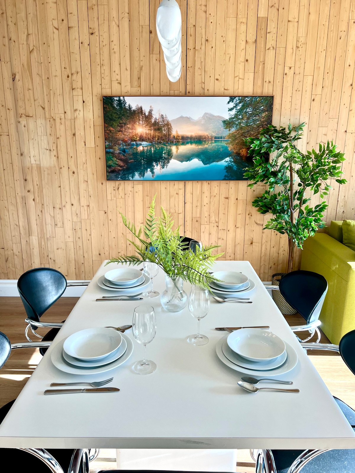 A dining table is set for a meal, featuring white plates and glassware. A decorative vase with greenery is centered on the table. The backdrop includes a wooden wall and a large landscape photograph showcasing a lake scene, with a nearby green seating area visible.