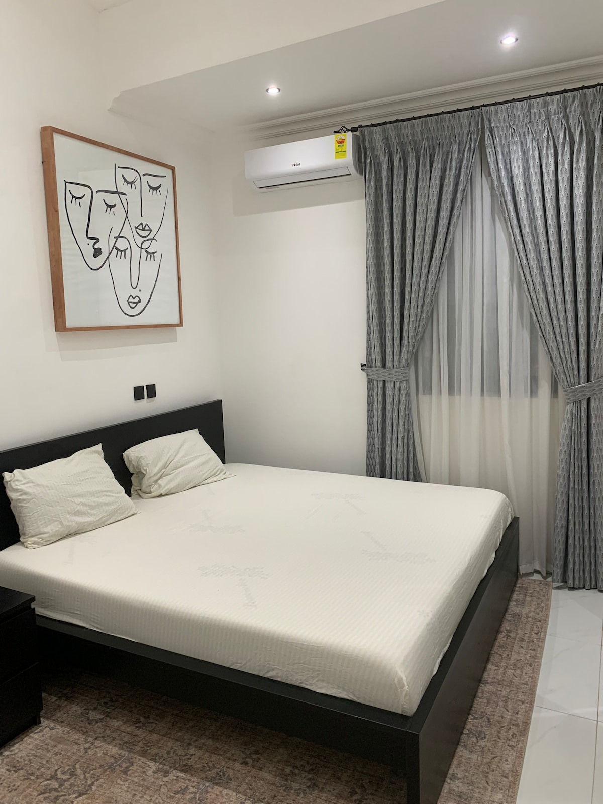 A serene bedroom is presented, featuring a spacious bed with neutral bedding against a light-colored wall. Large curtains frame a window, allowing natural light to softly illuminate the room. An abstract artwork is displayed above the bed, adding a touch of character.
