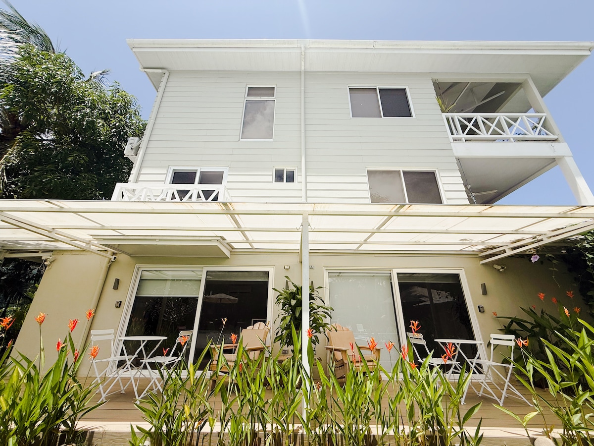 A two-story bungalow complex is displayed, featuring white siding and multiple large windows. The ground floor has a patio area adorned with tropical plants and vibrant flowers. Two white folding chairs are visible on the patio, inviting relaxation by the lush greenery.