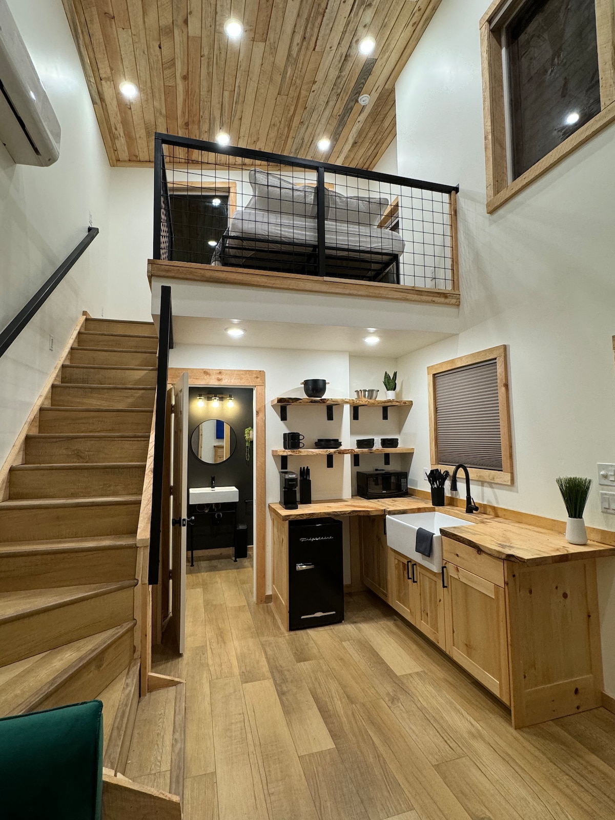A compact interior space is featured, showcasing a wooden staircase leading to a lofted sleeping area. The kitchenette includes a small refrigerator and a sink, with a stylish countertop offering necessary amenities. Natural light enters through a nearby window, illuminating the wood-paneled ceiling.