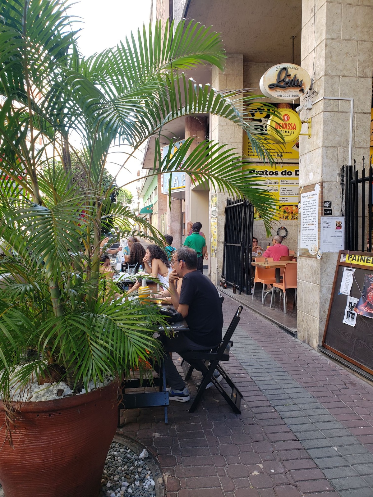A lively street scene features diners enjoying their meals at outdoor tables along a bustling sidewalk. Lush green palm leaves provide a natural border, while various storefronts, including a restaurant, contribute to the vibrant atmosphere of the area.