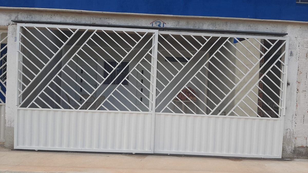 A white metal gate with diagonal patterns is shown, featuring a dual-panel design that opens to a driveway. The surrounding area is paved and a wall painted in blue can be seen in the background.