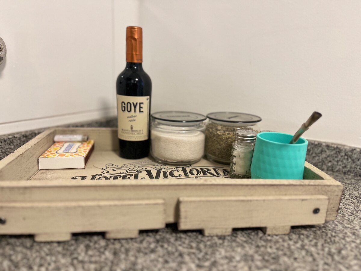 A wooden tray is arranged with a bottle of wine, two jars filled with spices, a sugar container, and a small turquoise cup. Two packets, presumably of snacks, are also included, all placed on a textured countertop.