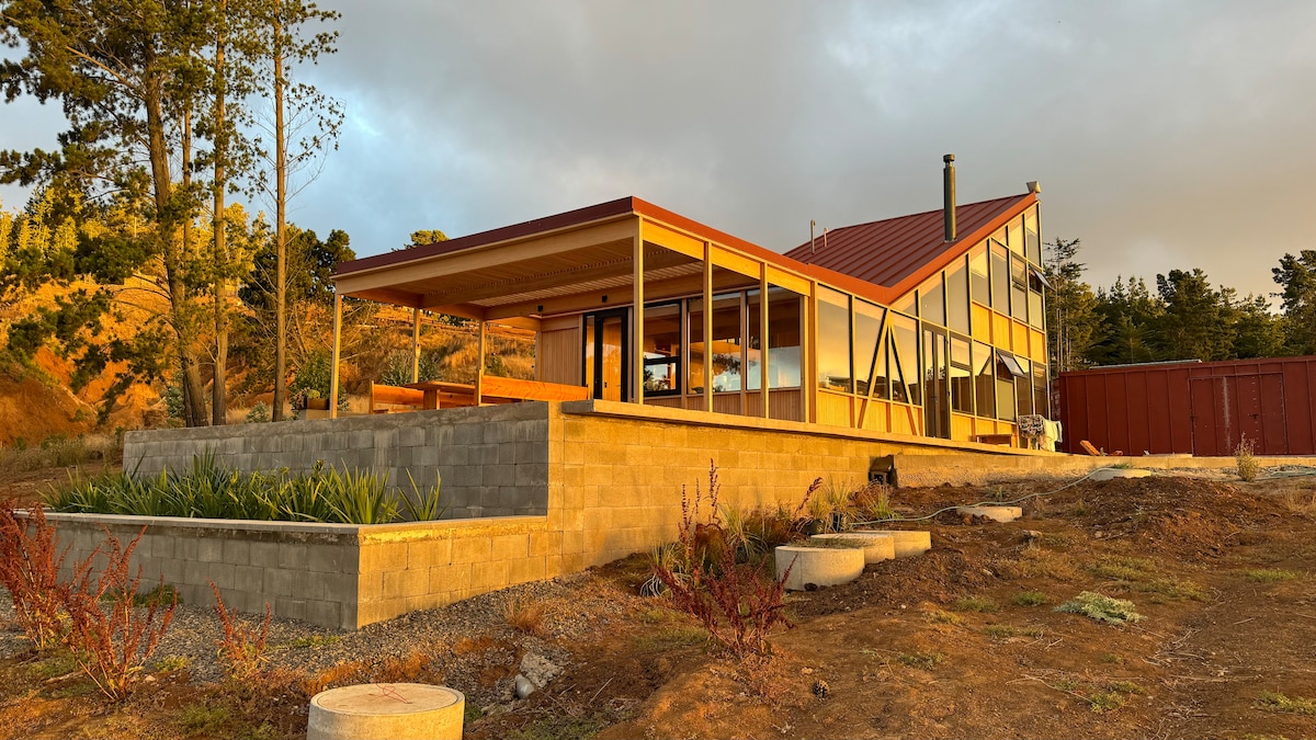 A modern house is presented with large windows and a sloped roof, showcasing a blend of wood and metal elements. Surrounding landscaping features low stone walls and patches of greenery, while the setting sun casts a warm glow over the structure, enhancing its architectural design.