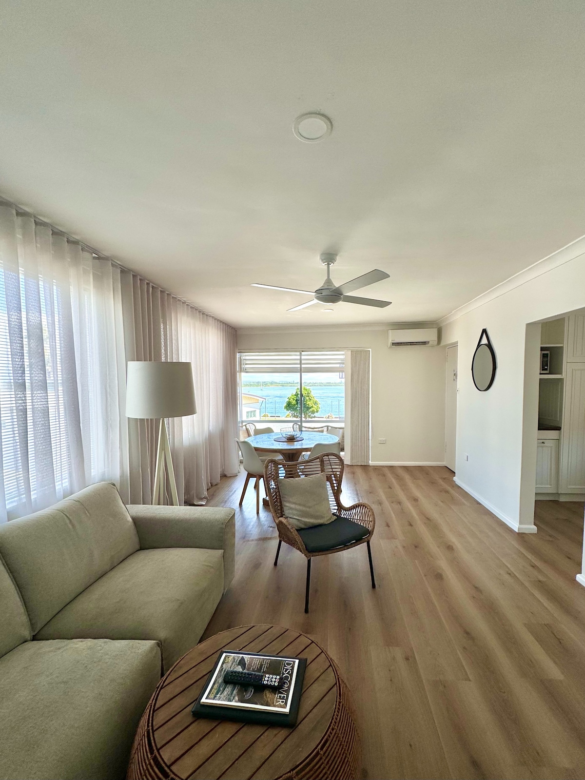 An open-plan living area is presented, featuring a comfortable beige couch adjacent to a round wooden coffee table. A ceiling fan hangs above, and natural light filters through sheer curtains at a large window, providing a glimpse of the outdoor view.