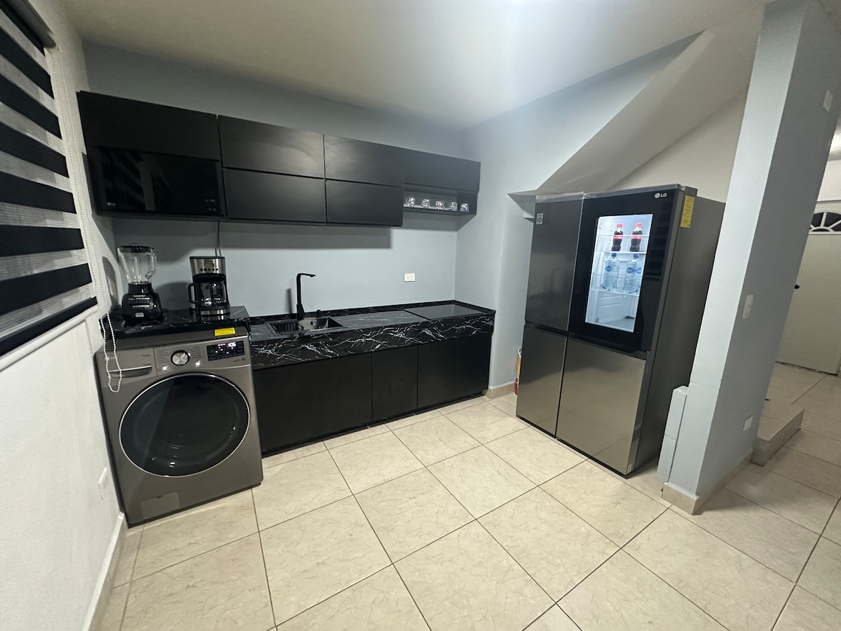 A modern kitchen is displayed, featuring sleek black cabinetry and a dark countertop. Appliances include a refrigerator, a washing machine, a coffee maker, and a blender. Neutral tile flooring is visible, alongside a window covered with a striped blind.