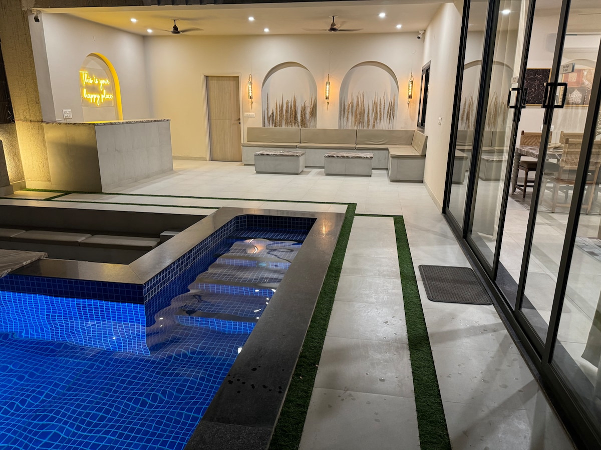 A private swimming pool is framed by sleek concrete, featuring a vibrant blue tile lining. Soft lighting enhances the modern space, while a seating area is situated nearby, offering a relaxed ambiance. Large glass doors provide access to a well-lit interior, inviting natural light.