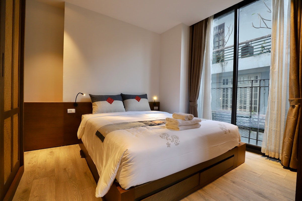 A well-lit sleeping area is presented, featuring a spacious bed adorned with decorative pillows. Soft linens cover the bed, flanked by warm lighting on each side. Large windows provide natural light and a view of the exterior, with light curtains allowing for privacy.