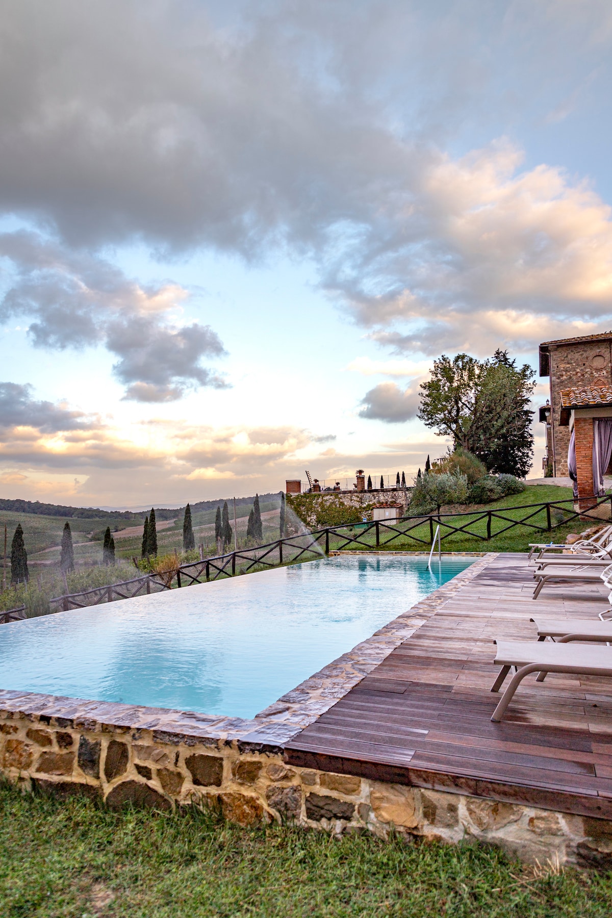 An infinity pool is situated against a picturesque backdrop of rolling hills and cypress trees. Light wooden sun loungers are arranged along the pool's edge, inviting relaxation as clouds drift across the sky. The tranquil setting offers scenic views of the surrounding landscape.