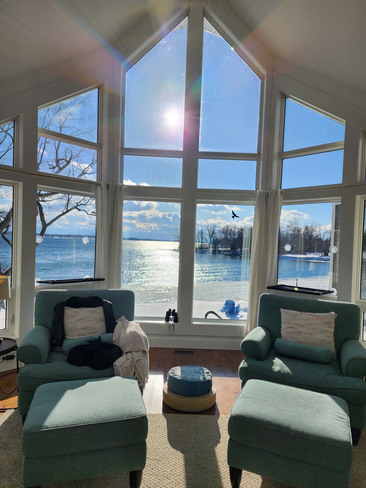 A bright living area features two comfortable armchairs positioned near large triangular windows overlooking the serene water. Sunlight reflects off the surface of the St Lawrence River, creating a tranquil ambiance. A cozy ottoman and a throw blanket are also visible, adding to the inviting setup.