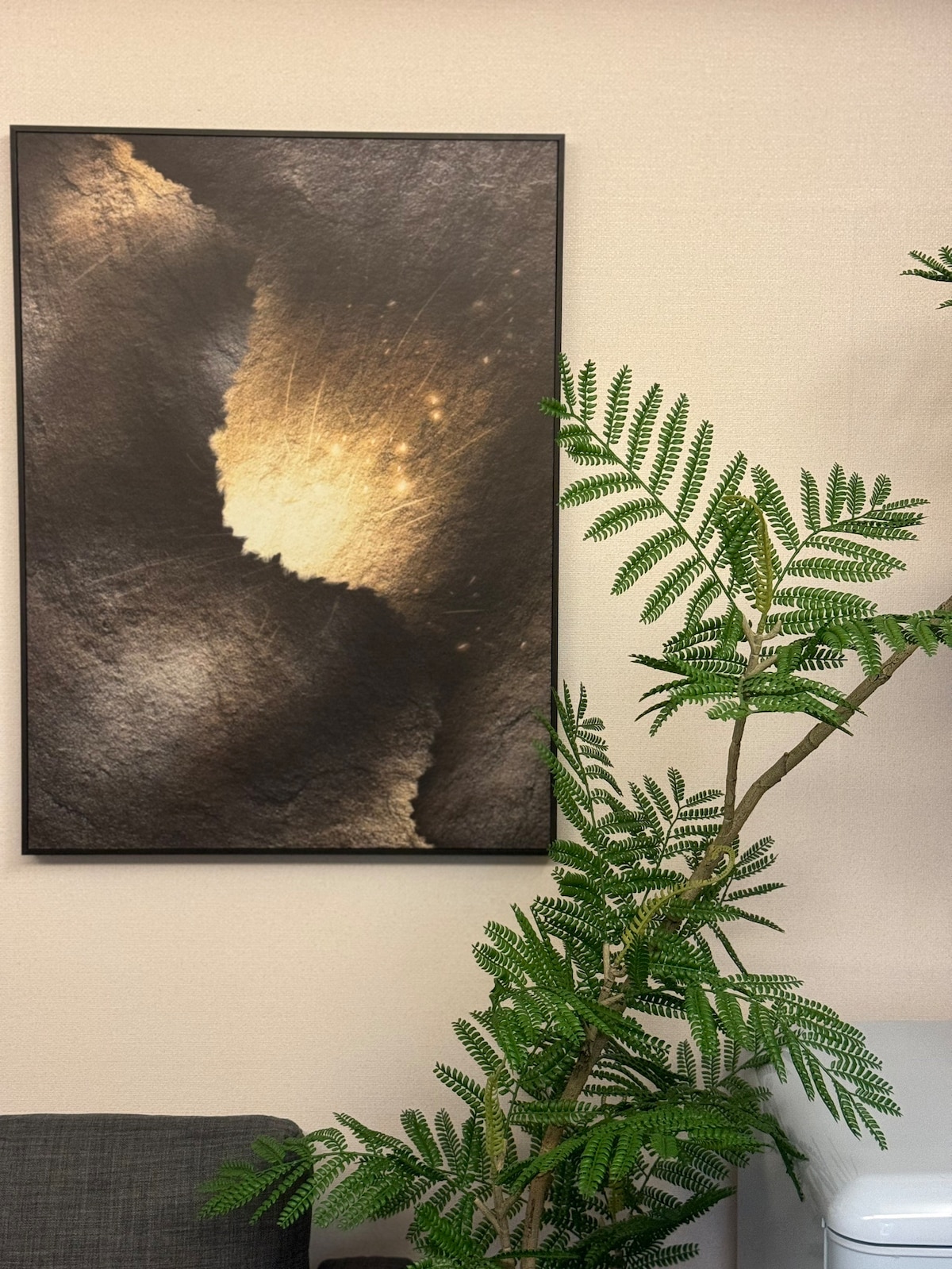 A framed artwork featuring abstract patterns in dark tones with hints of gold is displayed on a light wall. A green leafy plant is positioned nearby, adding a touch of nature to the space, while a soft gray sofa is partially visible in the foreground.