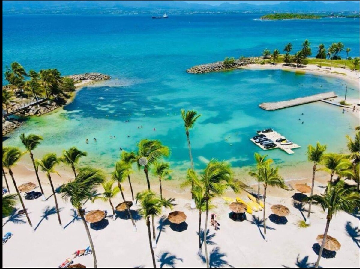 A scenic beachfront scene showcases clear turquoise waters gently lapping at a sandy shore, bordered by lush palm trees. Sunbeds and umbrellas are positioned along the beach, while small boats are moored nearby. The distant landscape features rolling hills under a bright blue sky.