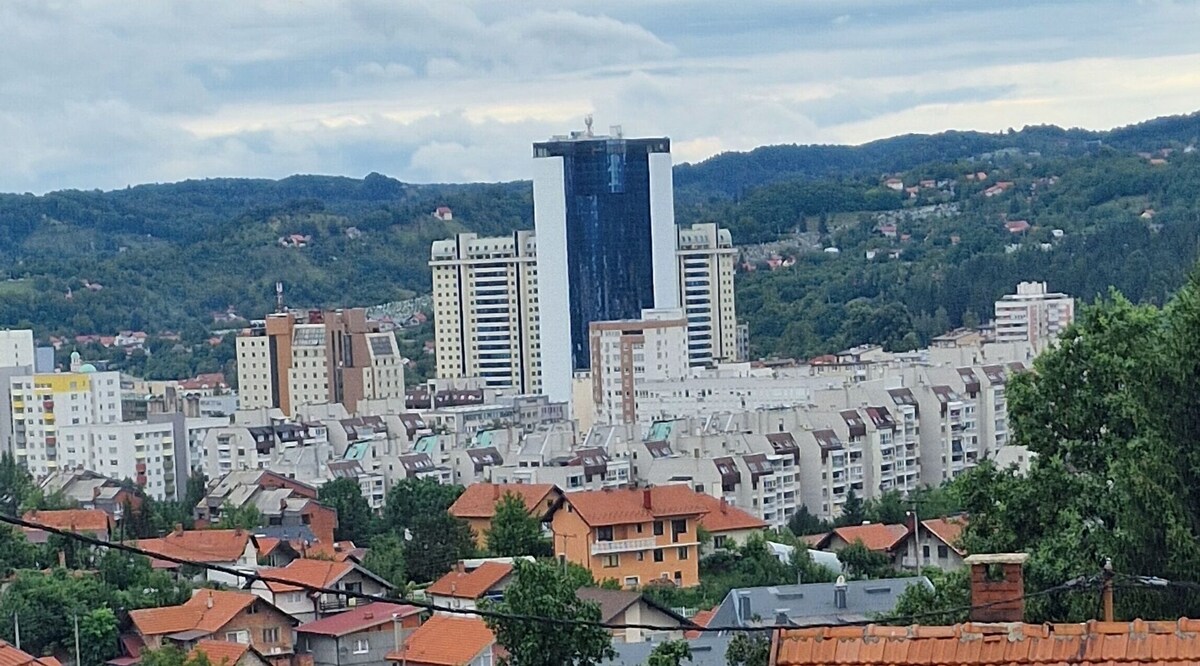 A panoramic view of Tuzla is presented, showcasing a blend of modern high-rise buildings and residential structures. Nestled among the hills, the rooftops display a mix of colors, with greenery in the background adding to the landscape.