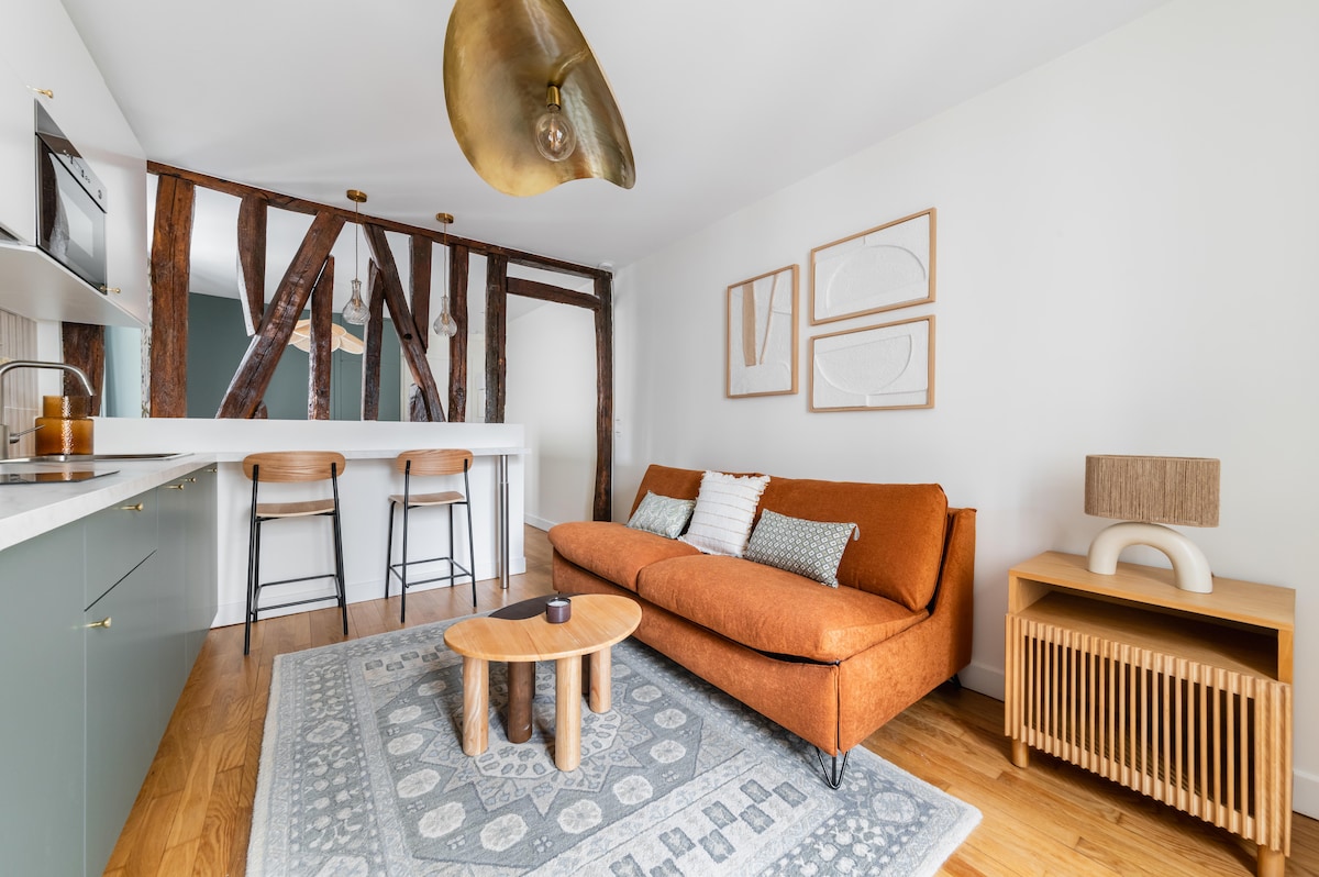 A modern living area features a comfortable orange sofa, complemented by a decorative rug beneath a wooden coffee table. Exposed beams add character, while bar stools sit at a kitchen counter in the background. Soft lighting is provided by an overhead fixture.
