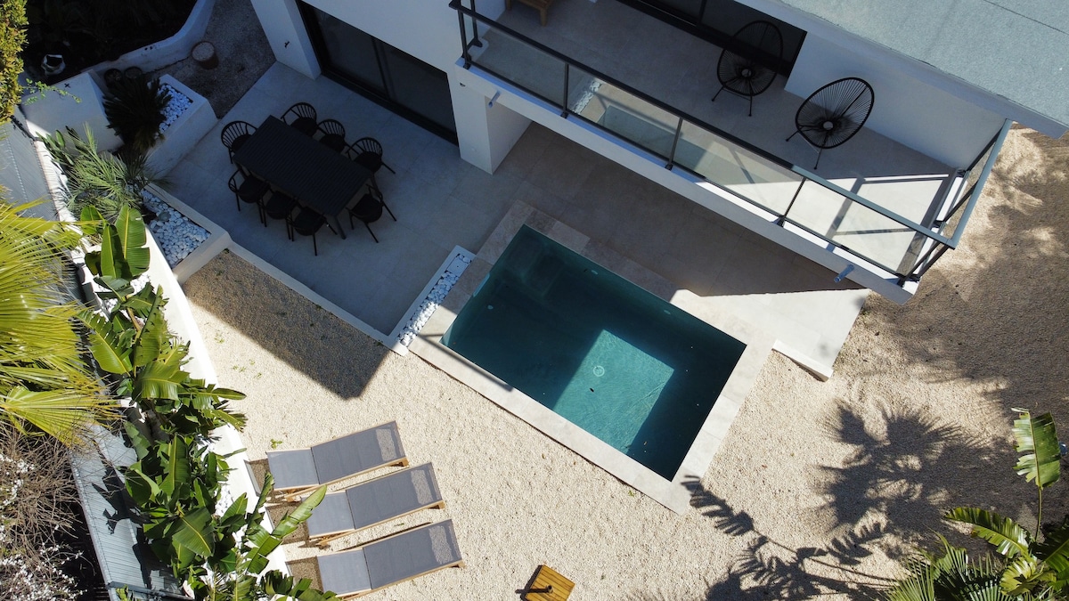 An aerial view of a modern villa's outdoor space features a small pool surrounded by a sandy area. A dining table with black chairs is positioned on the patio, alongside two lounge chairs for relaxation. Lush greenery is visible, enhancing the tranquil setting.