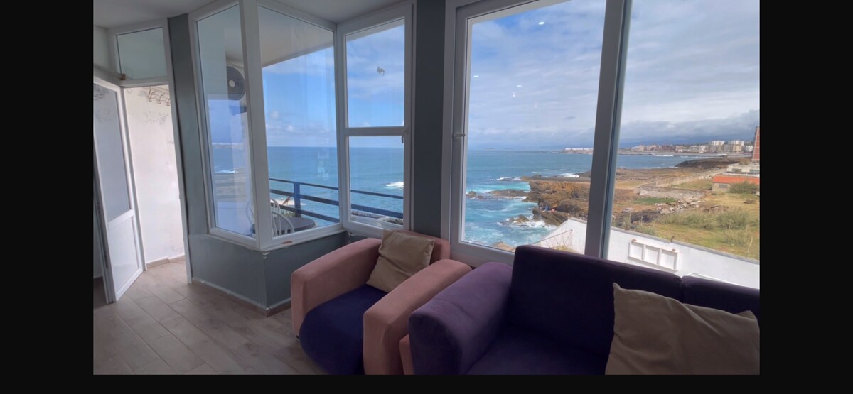 A seating area is highlighted by large windows providing panoramic views of the rocky coastline and waves. Two sofas, in contrasting colors, are positioned for comfort. Natural light fills the space, enhancing the serene atmosphere of the room by the sea.