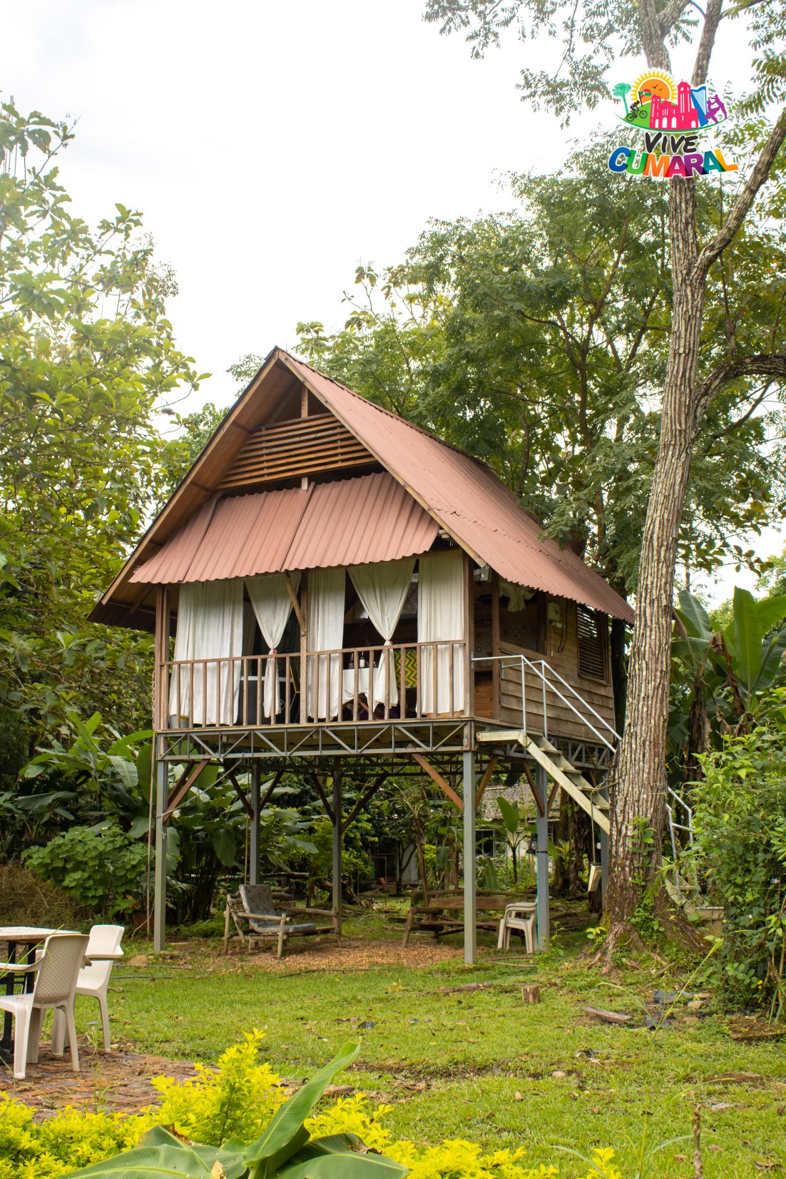 A charming elevated cabin is surrounded by lush greenery, supported by a sturdy wooden structure. The sloped roof is topped with metal sheeting. Several white curtains frame the windows, which provide a bright view of the outdoor setting. A small seating area is visible below.
