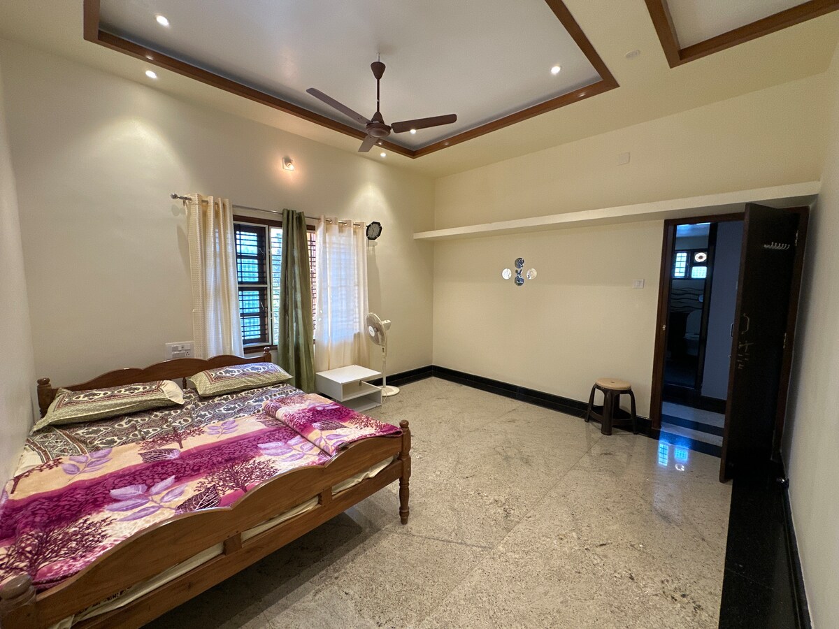 A spacious bedroom is furnished with a wooden bed dressed in a colorful coverlet. A fan hangs from the ceiling, while natural light filters through sheer curtains. A small stool is placed against the wall, and a doorway leads to another room, offering a seamless flow within the space.