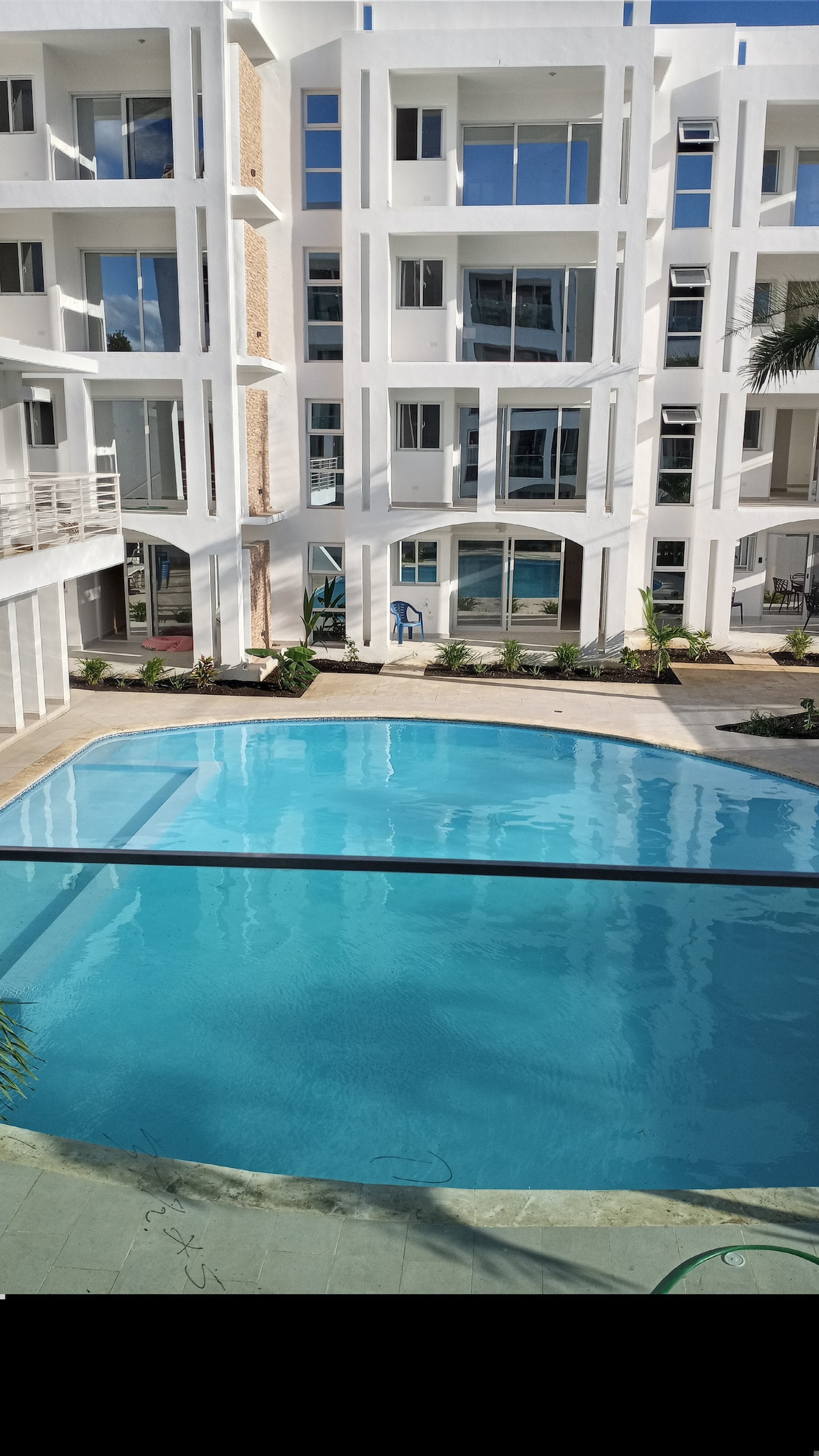 A bright, inviting pool is featured at the center of the urban complex, surrounded by modern white buildings. The water is clear and reflects the sky, while landscaped greenery has been incorporated around the pool area, enhancing the tranquil setting.