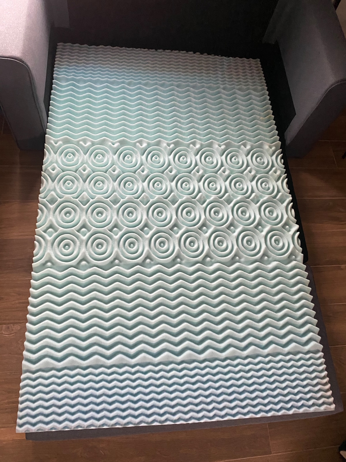 An innovative foam mattress topper is displayed on a textured surface. Its wavy and circular patterns are designed to enhance comfort. The topper's light blue and white colors create a soothing visual, complementing the surrounding furniture in the cozy space.