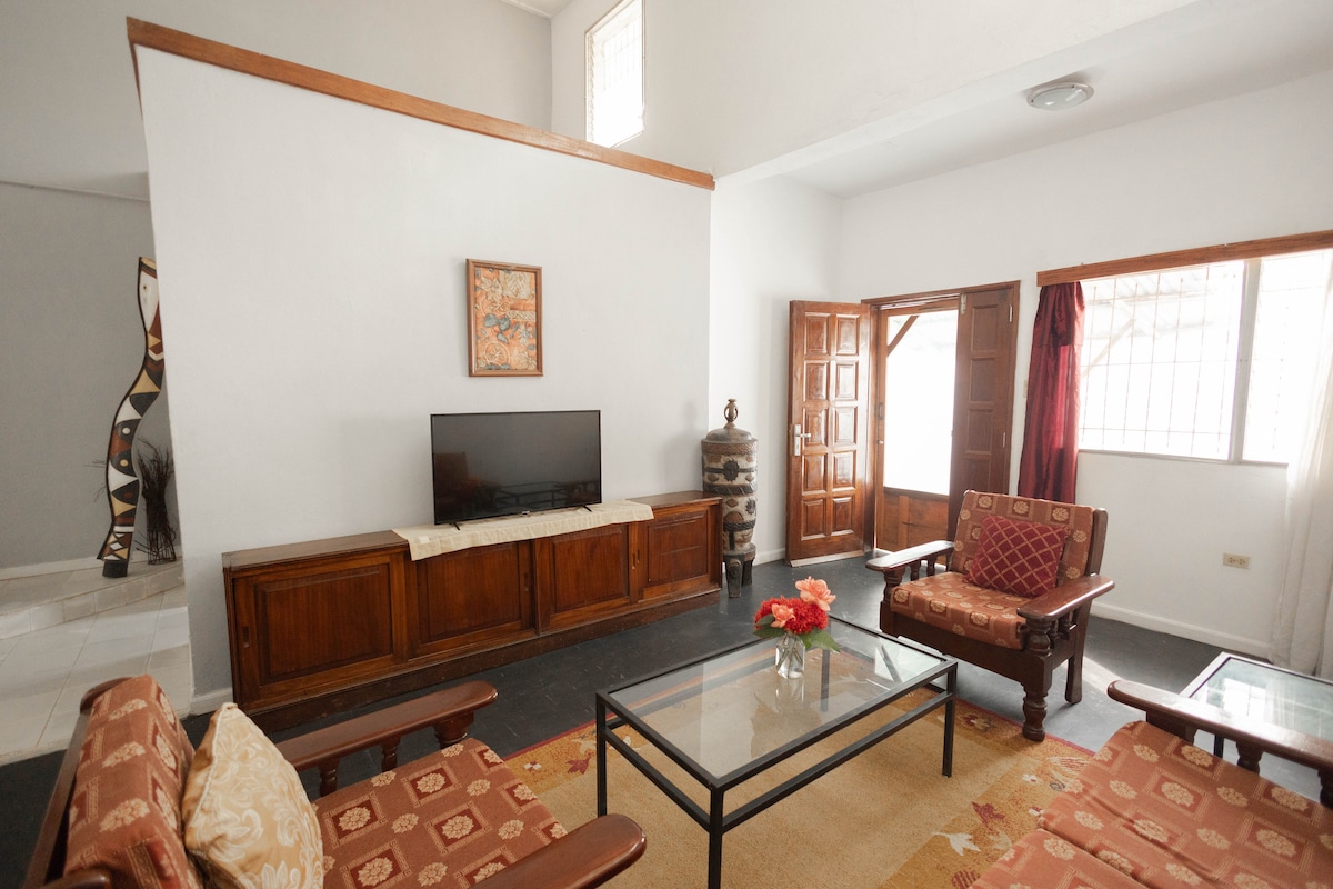 A living area features a large wooden cabinet and a flat-screen television mounted on the wall. Two wooden chairs with decorative cushions and a glass coffee table are positioned centrally. Natural light enters through the door and window, creating a bright atmosphere.
