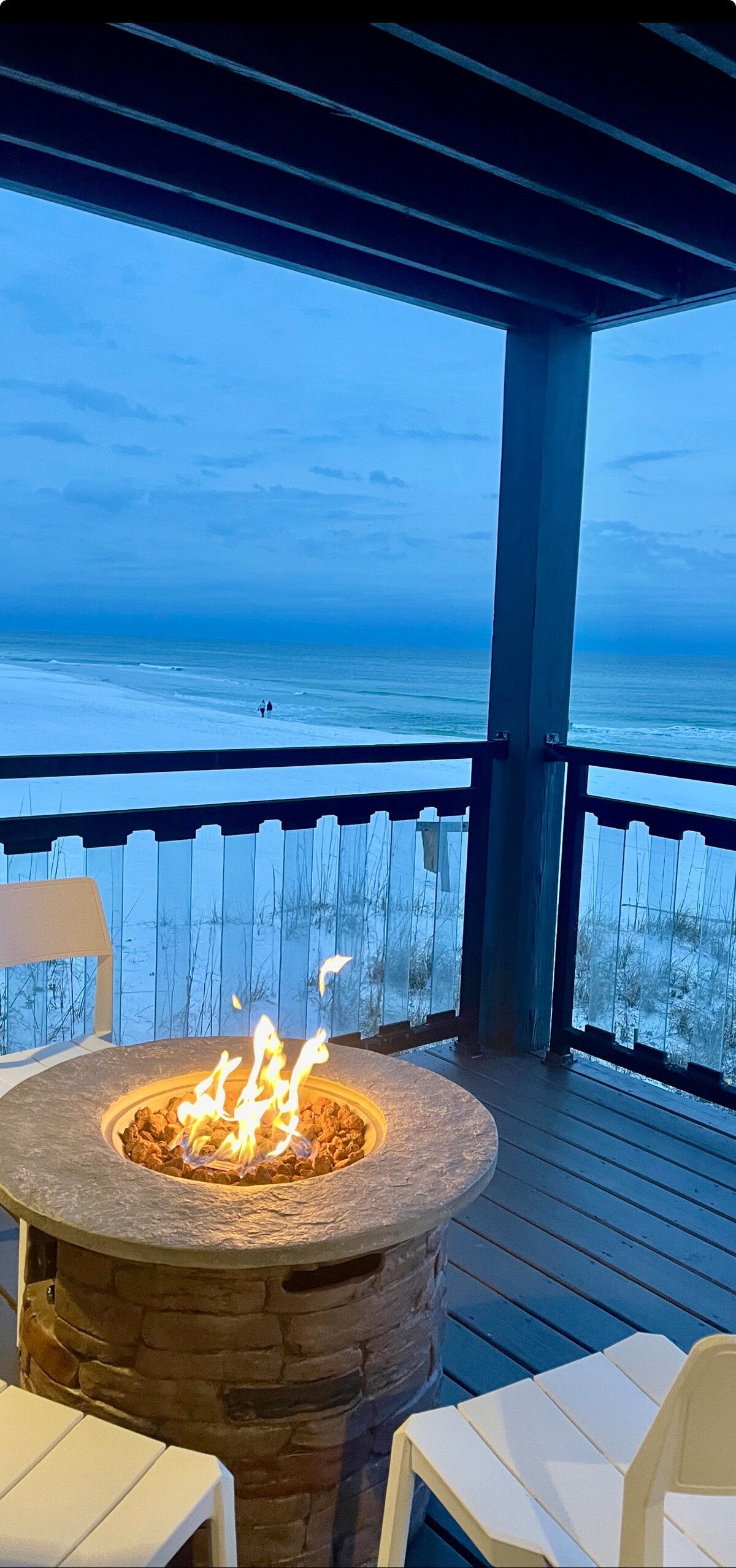 An outdoor seating area features a circular stone fire pit with flickering flames, surrounded by simple white chairs. The background showcases a serene view of the beach and ocean, with soft waves meeting the sandy shore under a twilight sky.