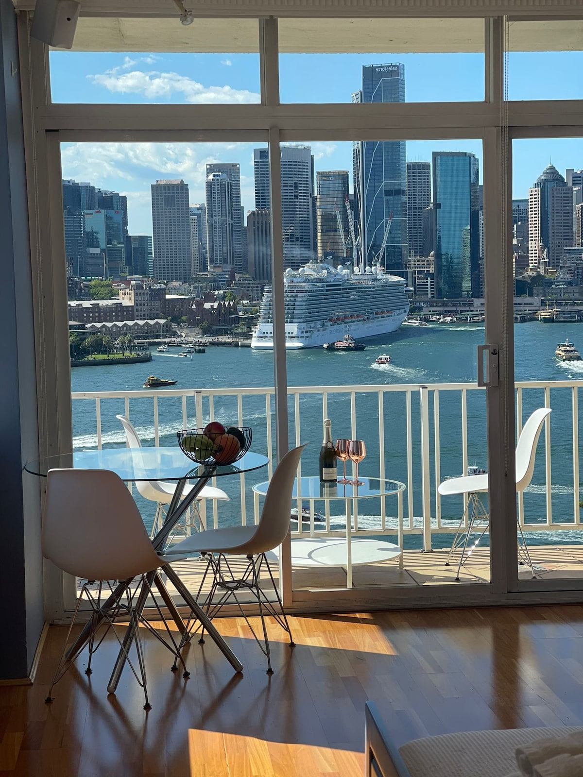 A balcony view captures the Sydney Harbour, featuring a bustling scene with a large cruise ship and the city skyline in the background. A small round table with white chairs is visible, with sunlight reflecting off the water.