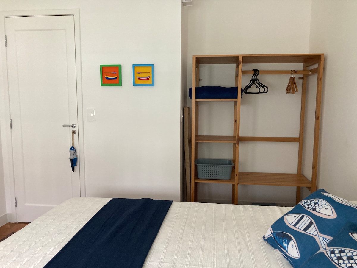 A cozy sleeping area features a spacious bed adorned with a decorative pillow. A wooden shelving unit holds hangers and storage bins. Two colorful art pieces hang on the wall, complementing the room's neutral tones. Light fills the space from a nearby window.