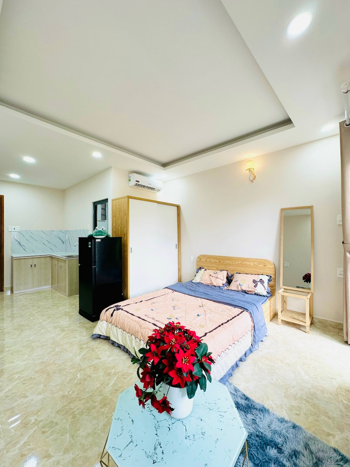 A well-lit bedroom features a spacious bed adorned with simple linens and decorative pillows. A mirror and a side table provide functionality. A small kitchenette is visible in the background, complemented by a sleek black refrigerator. A vase with flowers adds a touch of color to the room.