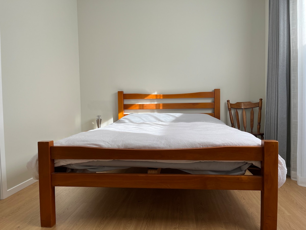 A wooden bed frame supports a spacious mattress, covered with a light grey blanket. A wooden chair is positioned beside the bed. Natural light filters through a curtain, enhancing the soft tones of the room.