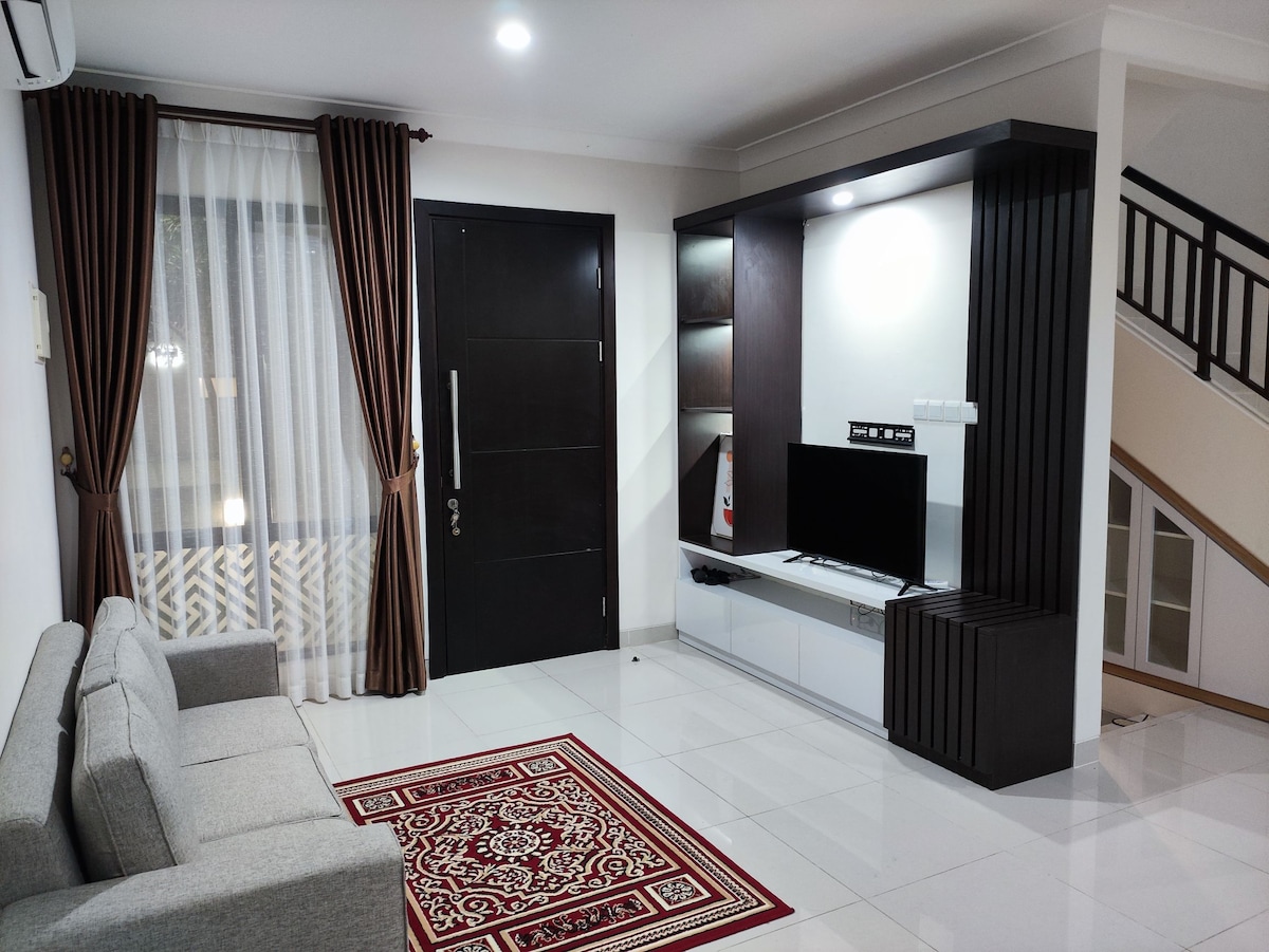 A modern living area features a comfortable gray sofa positioned beside a decorative rug. A wall-mounted television is placed on a sleek console, accompanied by elegant shelving. Natural light filters through a curtained window, illuminating the space.