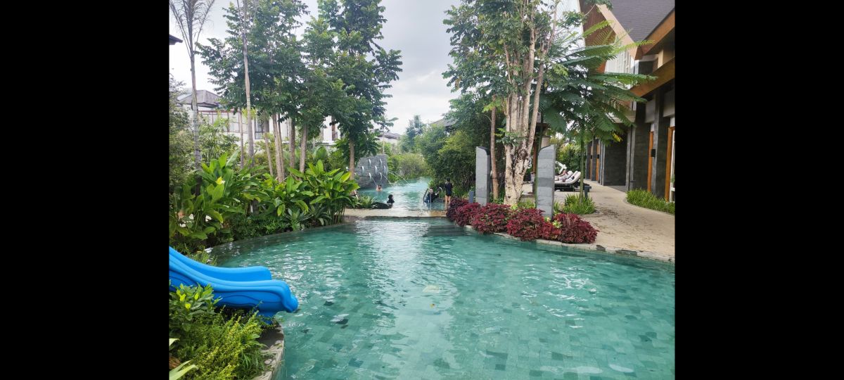 A tranquil swimming pool is surrounded by lush greenery, including tall trees and vibrant flowers. The clear blue water reflects the overcast sky, while a water feature adds a gentle sound. A slide is visible along the pool's edge, inviting leisure activities.