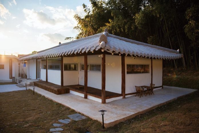 A spacious traditional-style house features a pristine white exterior with a tiled roof. Wooden beams support an inviting porch area, accompanied by a small outdoor table and chairs. Sunlight casts a warm glow on the surrounding grassy area, complemented by a backdrop of bamboo.