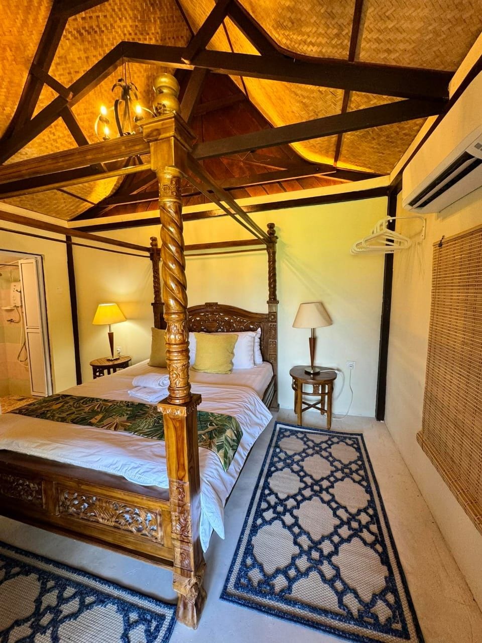 A wooden four-poster bed is presented with white linens and a decorative throw. Two lamps provide soft lighting beside the bed. A patterned rug lies on the floor, and natural materials enhance the ceiling's design, creating a serene atmosphere.