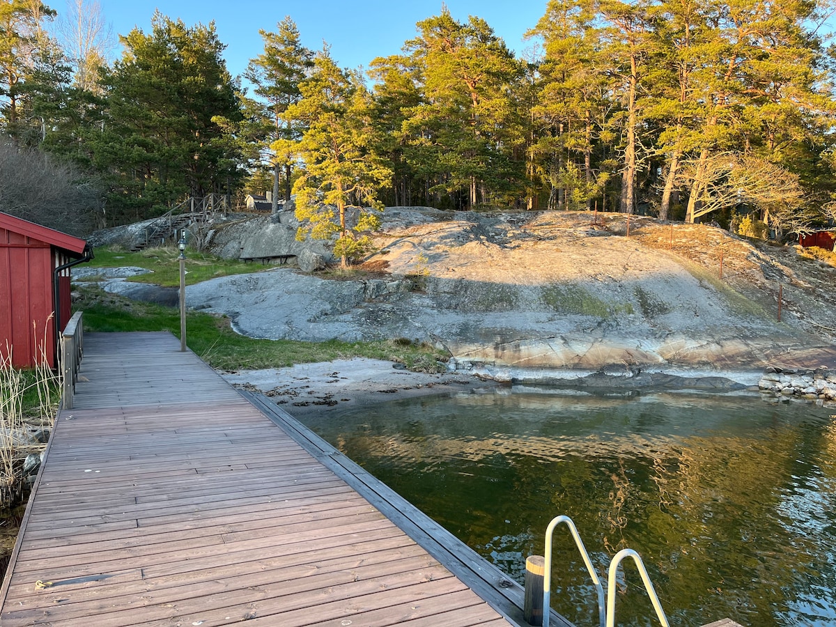A wooden deck leads to the shoreline, surrounded by natural rock formations and tall trees. The calm water reflects the blue sky and distant greenery, creating a serene setting. A set of steps provides access to the water for swimming.