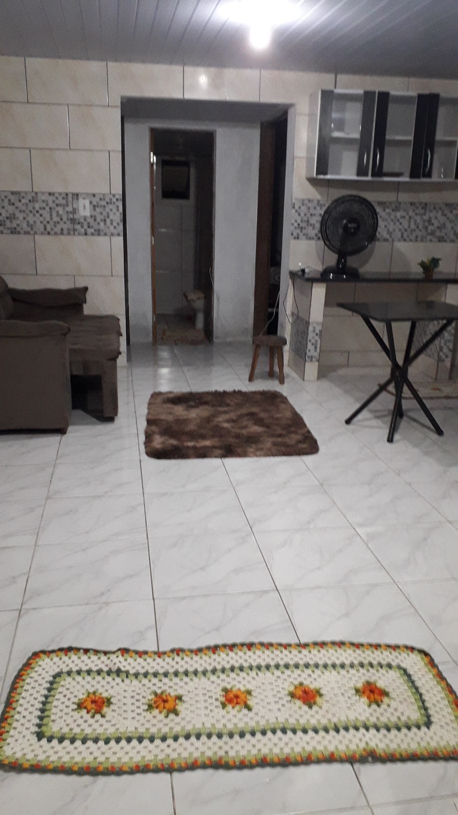A spacious living area is showcased, featuring a comfortable sofa and a small table with a fan. Two area rugs are placed on the tiled floor. Light-colored walls with a tiled backsplash provide a clean backdrop, and a doorway leads to another room.