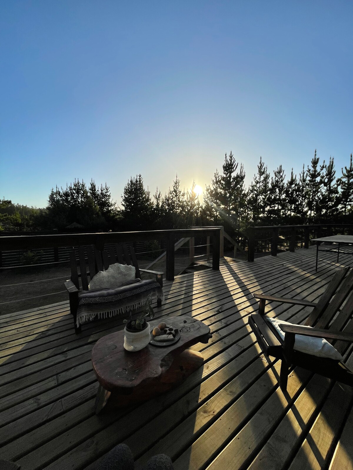 A spacious wooden deck is shown, featuring wooden furniture including a cozy chair and a rustic coffee table. The sun is rising in the background, casting soft light across the area, with tall trees framing the view.