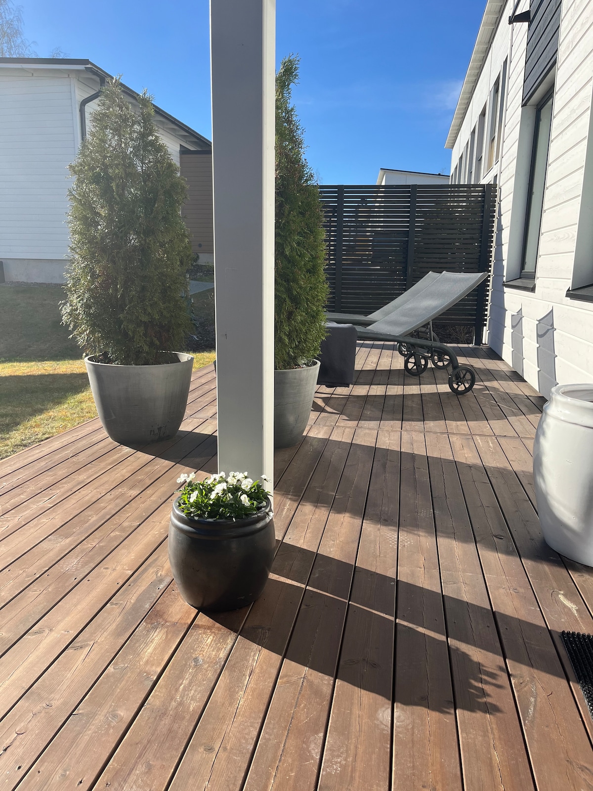 An outdoor terrace features a wooden deck with potted plants, including a flowering arrangement. A reclining chair is positioned nearby, providing a spot to relax. Tall evergreen plants provide a sense of privacy, complemented by bright sunlight illuminating the space.