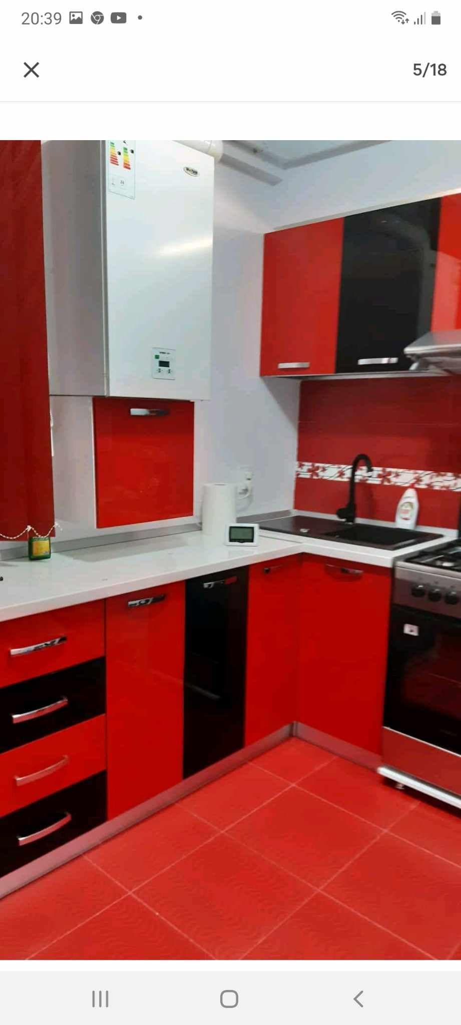 A kitchen featuring a modern design with red and black cabinets, complemented by a gray countertop. Appliances include a gas stove and a hood, along with a sleek black faucet. The floor is tiled in red, and a small window allows natural light to enter.