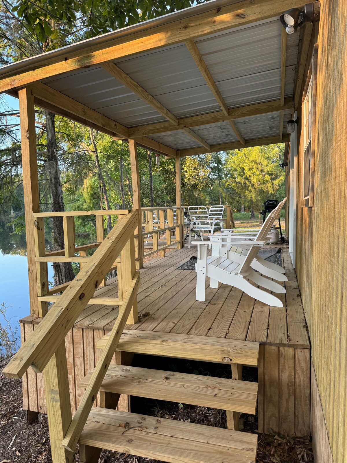 A partially covered wooden deck features several white Adirondack chairs arranged for comfort. Wooden steps lead down to the ground, with a scenic view of McDowell Lake visible in the background surrounded by lush greenery.