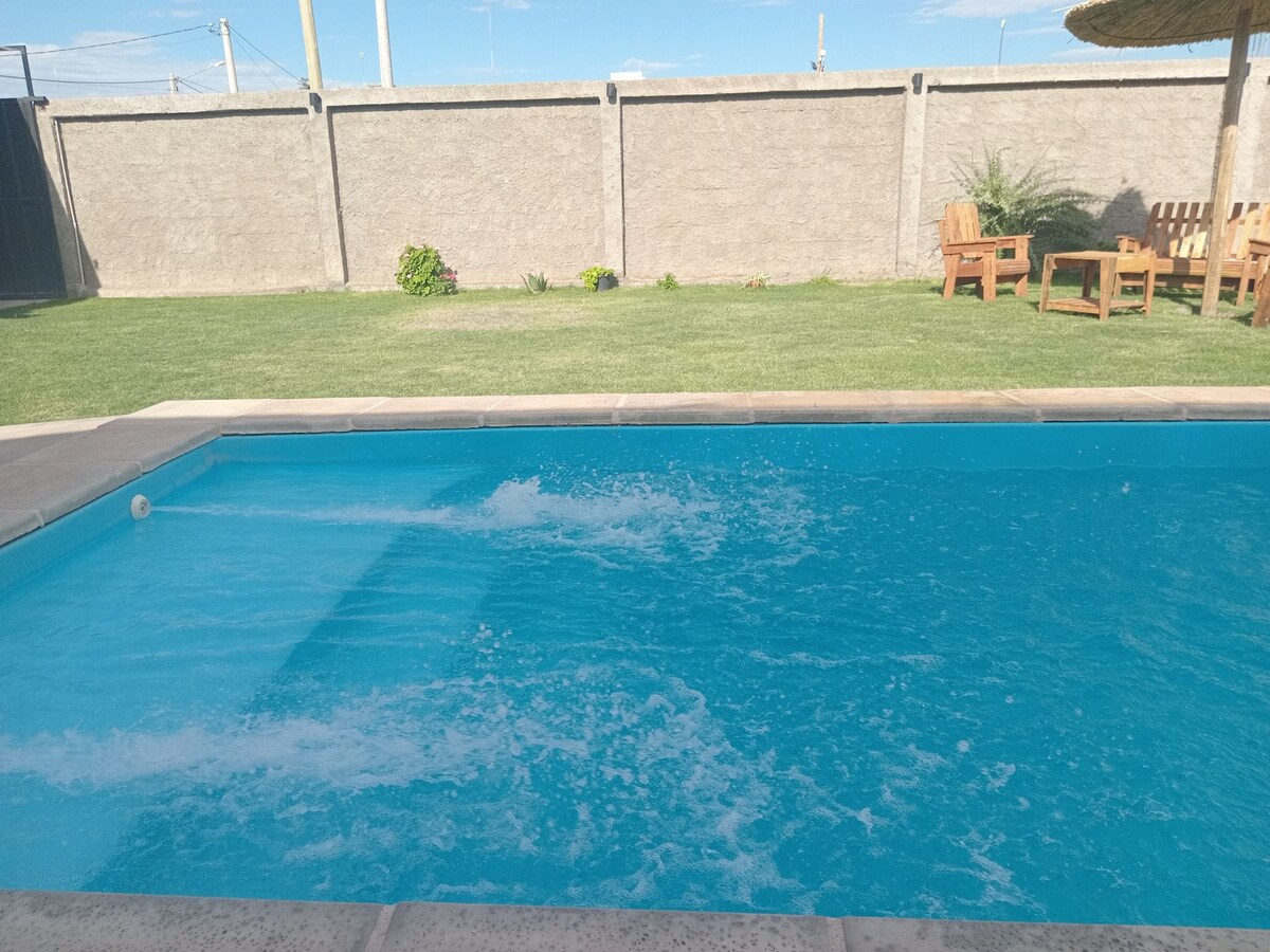 A clear swimming pool is surrounded by a well-maintained lawn. Wood chairs and a table are positioned nearby under a shaded structure. The pool's surface reflects sunlight, creating a vibrant blue hue.