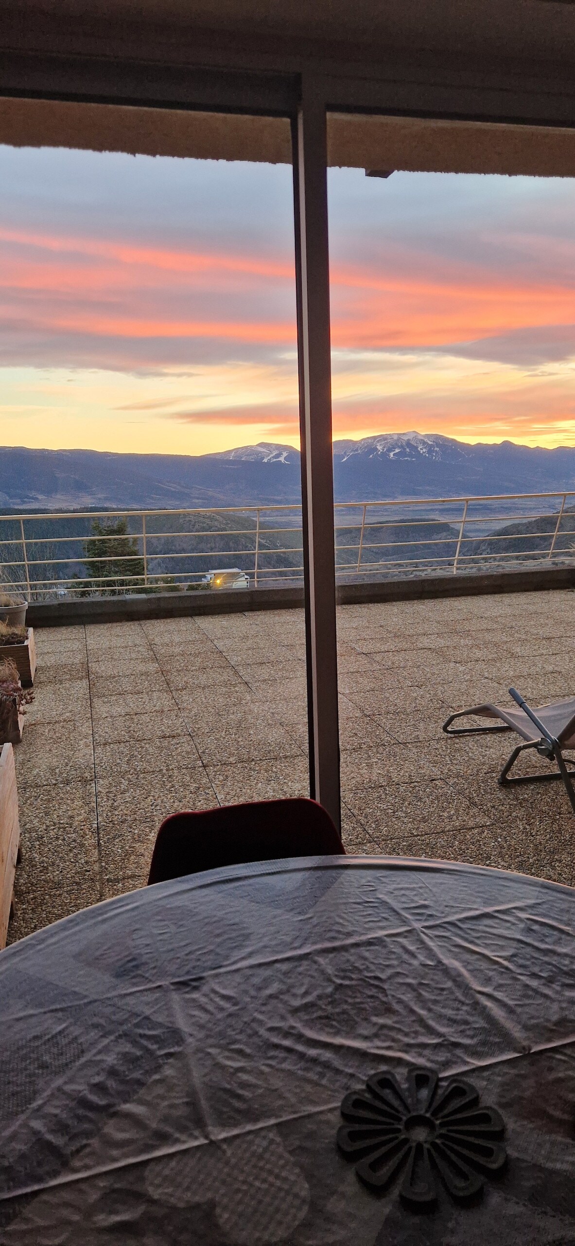 A spacious terrace features a table set against a backdrop of vibrant sunset colors. Snow-capped mountains rise in the distance, framed by large sliding doors that open to the outdoor space. The terrace offers ample room for enjoying the scenic views.