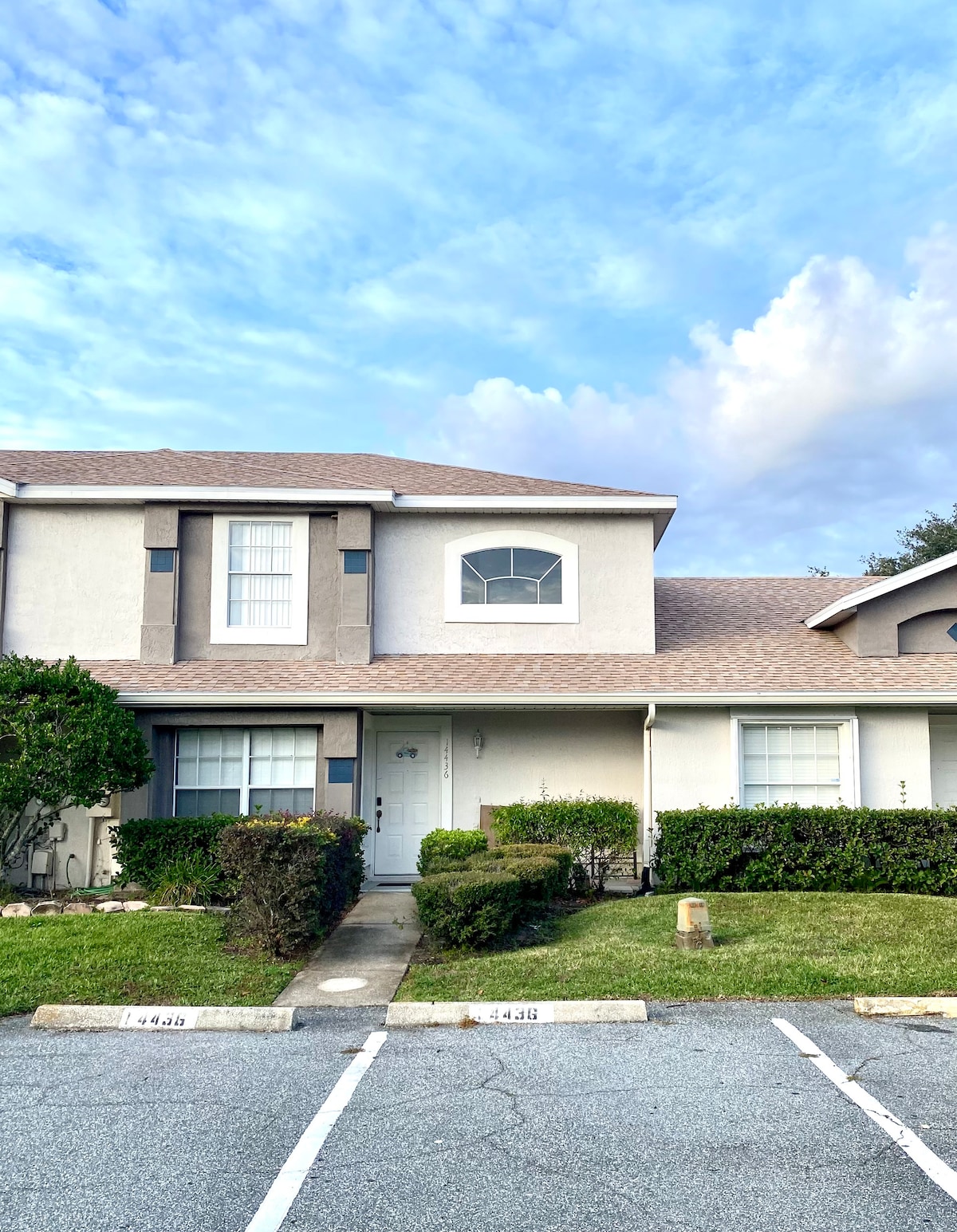 A two-story townhome is seen from the exterior, featuring a neutral color palette and symmetrical windows. Green shrubs and manicured lawns frame the entrance, while a paved walkway leads to the front door. The sky is partly cloudy, enhancing the inviting atmosphere of the property.