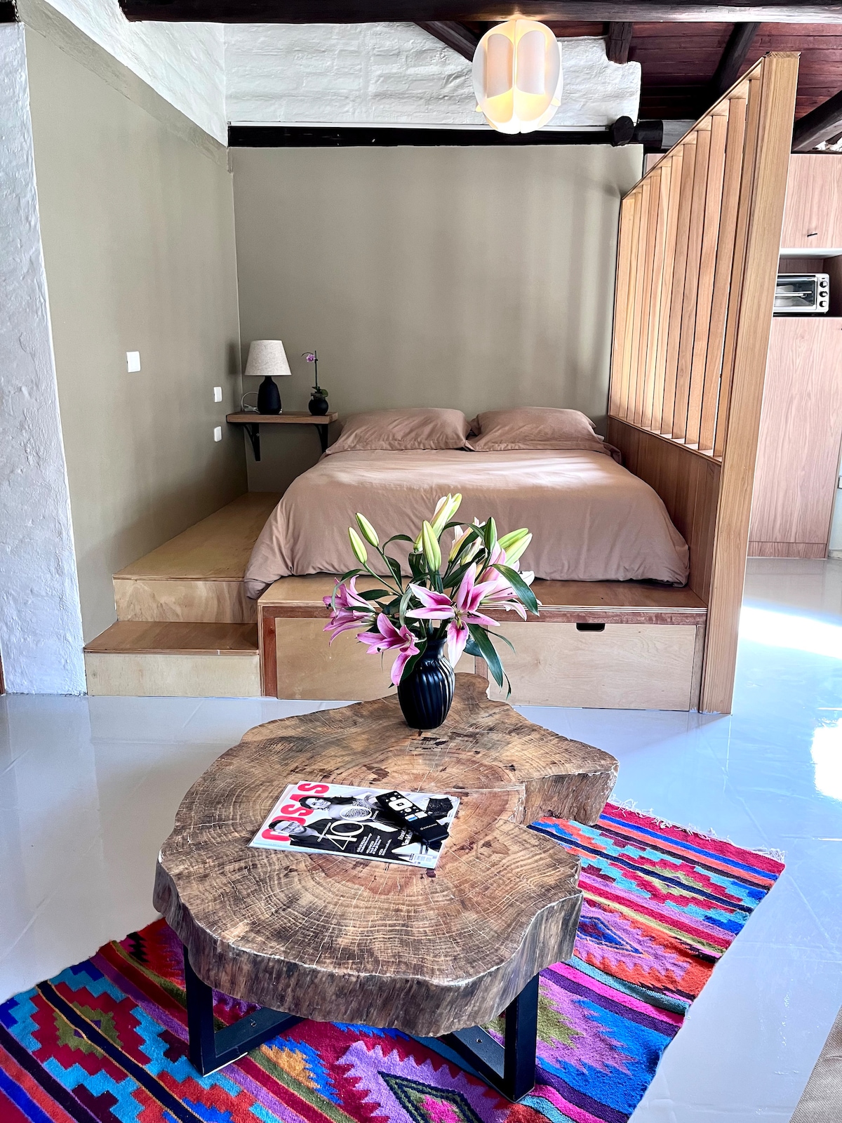A spacious sleeping area features a comfortable two-plaza bed with soft bedding, complemented by a bedside lamp. A unique wooden coffee table with a floral arrangement sits on a colorful area rug, enhancing the inviting ambiance of the studio.