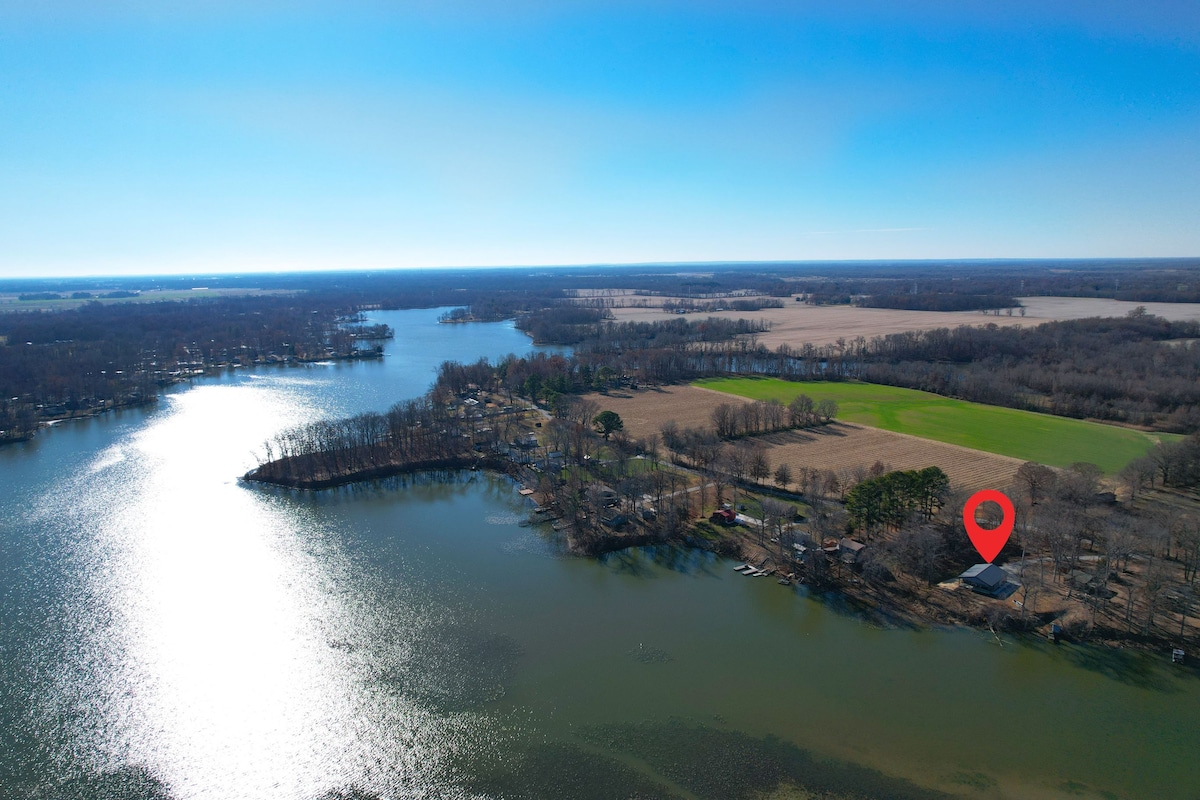 An expansive aerial view of a lakefront property is presented, surrounded by lush greenery and open fields. The lake sparkles under bright sunlight, while a red marker indicates the location of the home, highlighting its proximity to tranquil waters and scenic landscapes.