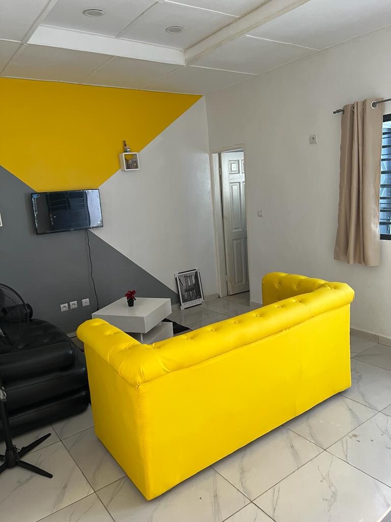 A bright yellow couch is positioned in a modern living space, complemented by a contrasting gray and white wall backdrop. A small white coffee table is situated in front, while a wall-mounted television is visible. Natural light enters through a window with light curtains.