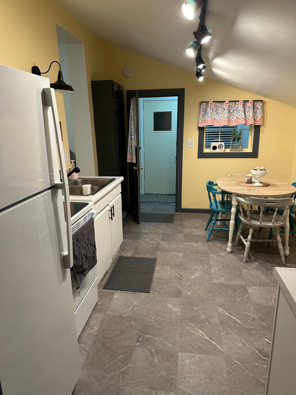A light-filled kitchen features a white refrigerator and a stove with a hood. A small dining table with colorful chairs is visible, along with cheerful curtains adorning the window. The flooring is tiled in neutral tones, enhancing the space's clean, modern design.