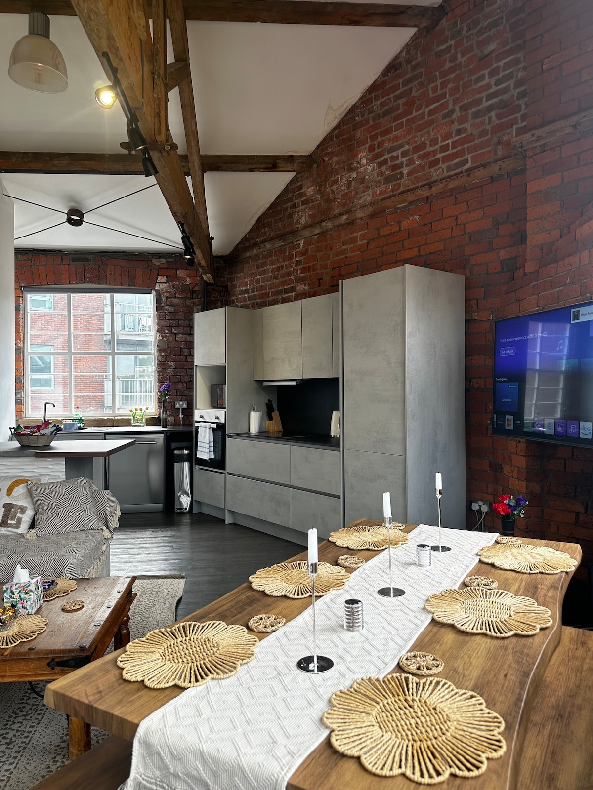 A dining area features a wooden table set with decorative woven placemats and candles. The background reveals a modern kitchen with gray cabinetry and stainless steel appliances, contrasted against the warm red brick walls and exposed wooden beams of the loft.