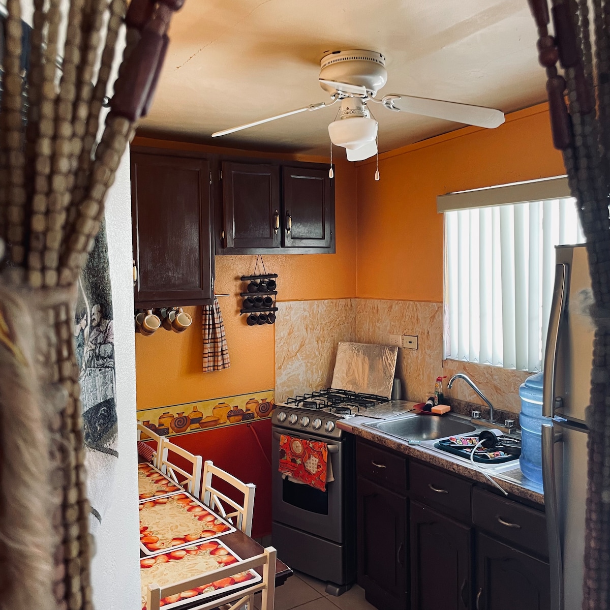 A functional kitchen is displayed with orange walls and dark wooden cabinetry. A gas stove is seen alongside a stainless steel refrigerator. A small dining table accommodates four chairs, and a ceiling fan is positioned above for airflow. Light filters through vertical blinds.